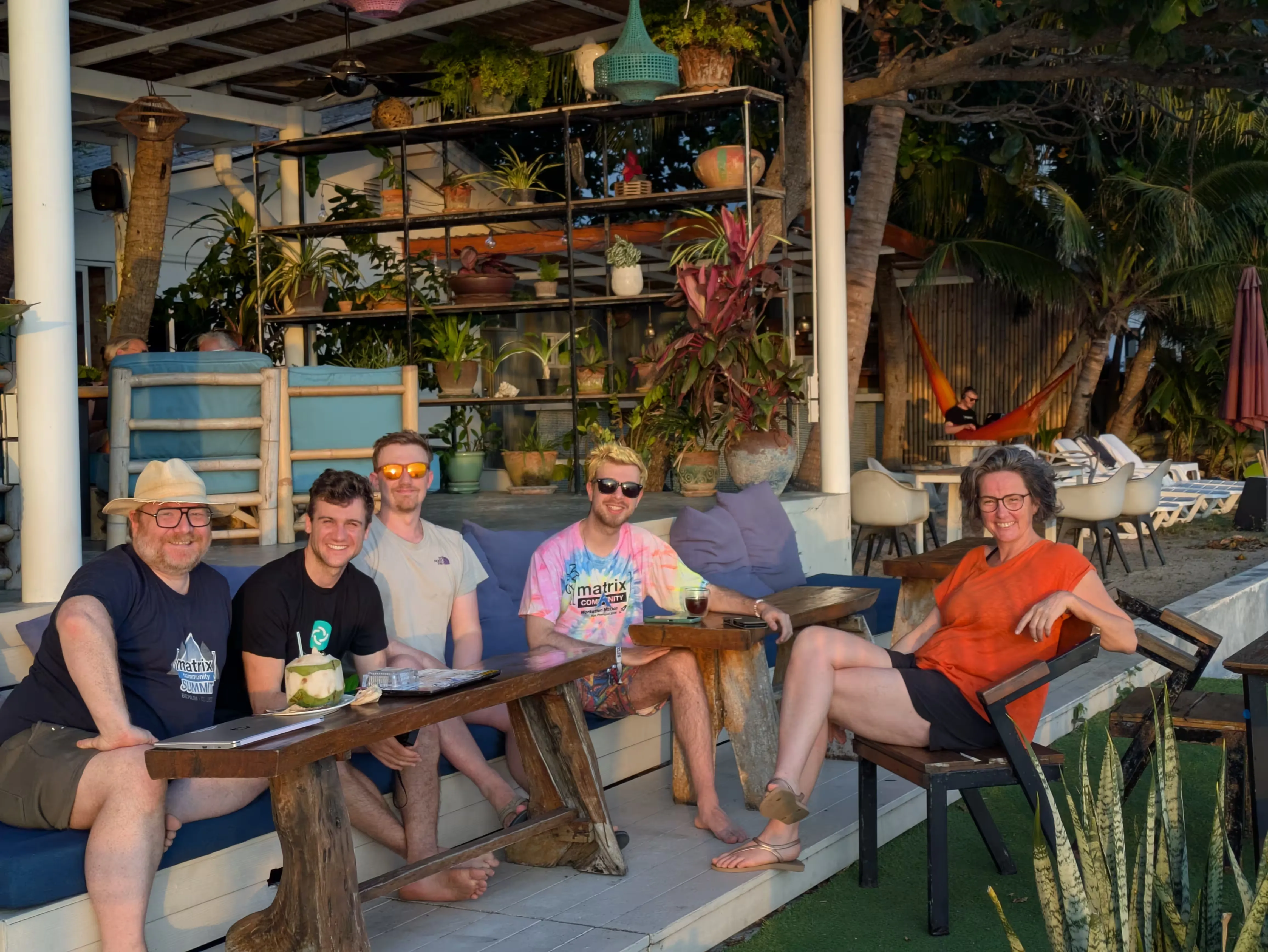 A group of people in summer outfits lounging on a sofa, enjoying the sunset. One is participating in a meeting from a hammock in the background.