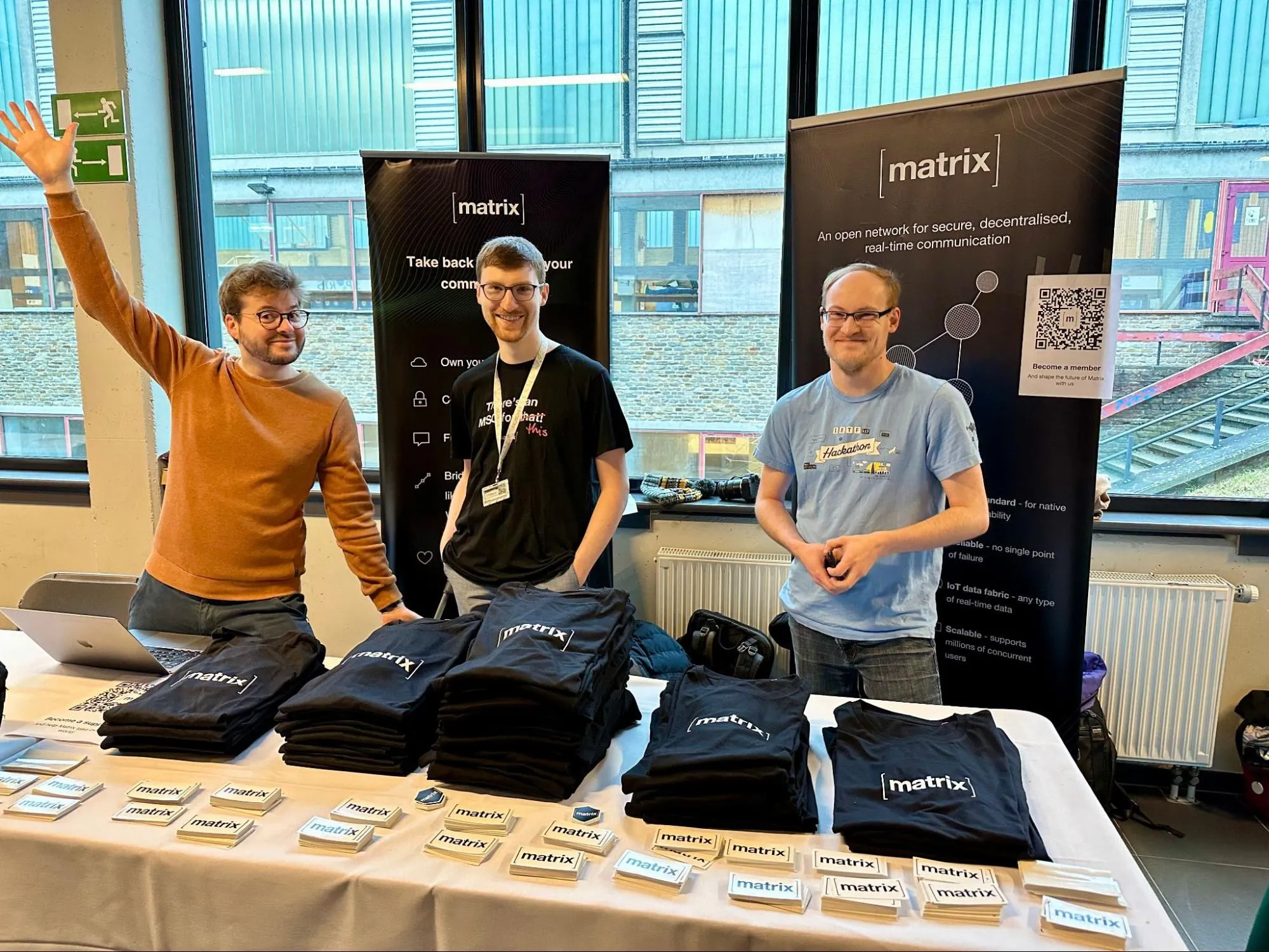 A picture of three persons standing behind a table. One of them has his arm stretched, the two others are looking at the camera smiling. There are folded t-shirts and stickers on the table.