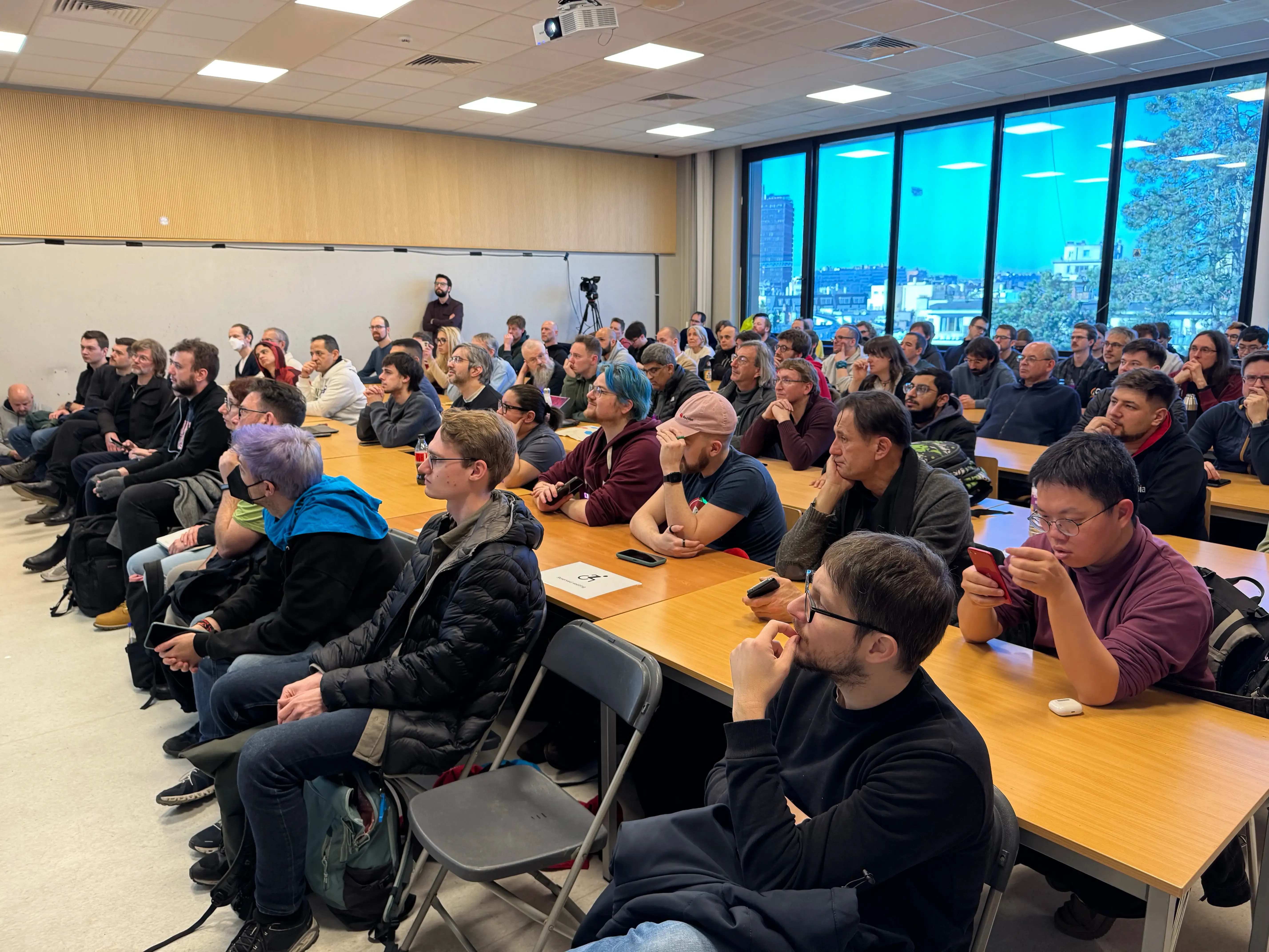 A picture of people sitting in a university lecture room. They are listening to a speaker off the frame. The room is almost full.